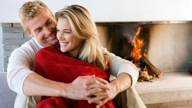 Imagem de um casal sentado próximo a uma lareira. Ele a abraça. Ela está com uma coberta na cor vermelha.