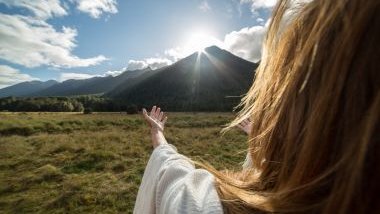 Imagem de uma mulher de costas olhando para um monte verde em frente ao Sol.
