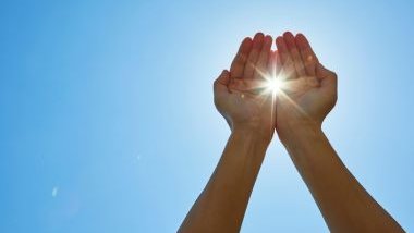 Imagem das mãos erguidas para o céu azul e no meio das mãos sai uma fresta da luz do sol.