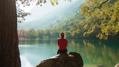Imagem de uma região de montanhas e árvores verdes com um lindo lago em destaque e uma mulher sentada em uma pedra observando a paisagem.