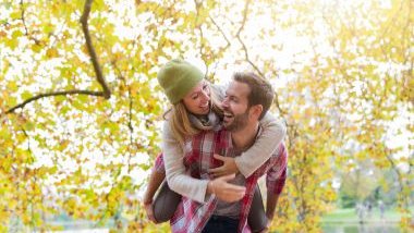 Imagem de um casal em uma praça. Ele está carregando a moça nas costas. Eles estão sorrindo e muito felizes.