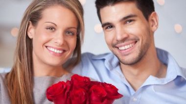 Imagem de um casal sorrindo e feliz. Ela segura em suas mãos, um lindo buquê de rosas vermelhas.