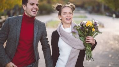 Imagem de um casal caminhando em um parque durante o inverno. Eles estão de mãos dadas e ela segura um buquê de flores do campo.