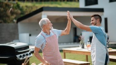 Imagem do sogro e seu genro fazendo churrasco na área gourmet. Ambos usam avental de cozinha, estão felizes e se cumprimentando com as mãos ao alto.