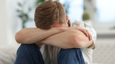 Imagem de um homem, sentado no sofá com o rosto debruçado sobre os seus braços. Ele está triste.