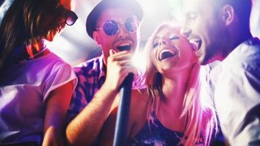 Imagem de quatro jovens, homens e mulheres cantando em um microfone, felizes e sorridentes.