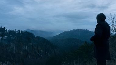 Imagem de uma montanha e uma floresta ao fundo. Do lado, a silhueta de um homem usando uma capa com capuz, simbolizando a entidade Zé do Mangue.