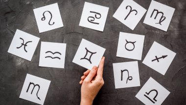 Cada símbolo de um signo do zodíaco está desenhado em um pedaço de papel. Os papéis estão espalhados em cima de um fundo cinza.