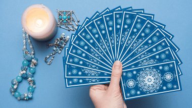 A mão de uma taróloga segurando diversas cartas de Tarot em sequência está em destaque na imagem. Ao lado, há uma vela e pingentes.