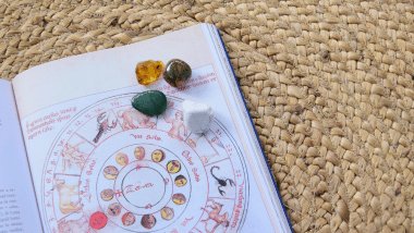 No foco da imagem, há um livro com uma Roda do Zodíaco desenhada e há pedras e cristais em cima do livro.