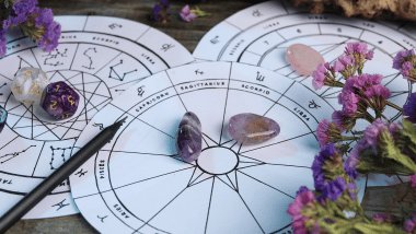 No foco da imagem, há três Rodas do Zodíaco desenhadas em papéis com os símbolos dos signos. Ao redor, há pedras e plantas.