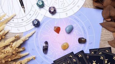 Há três rodas do zodíaco com os símbolos dos signos desenhadas em papel sulfite em cima de uma mesa. Ao redor, há cartas de Tarot e pedras.
