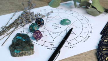 Há uma roda do zodíaco com os símbolos dos signos desenhada em papel sulfite em cima de uma mesa. Ao redor, há flores, pedras e dados.