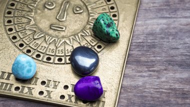 Pedras coloridas sobre placa com símbolo solar e signos gravados, apoiada em superfície de madeira.