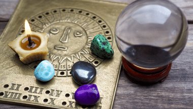 Bola de cristal, vela acesa e pedras coloridas sobre placa com símbolo solar e signos, em mesa de madeira.
