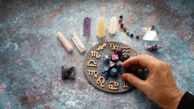 No foco da imagem, há uma Roda do Zodíaco com os símbolos dos signos e há dados, pedras e cristais ao redor.