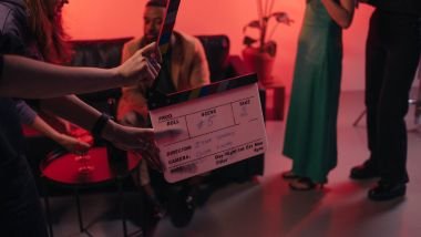 Imagem de atores em cena de um filme e em destaque o produtor segurando uma claquete durante a gravação.