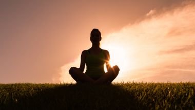 Imagem de uma mulher sentada em um gramado fazendo a sua meditação e ao fundo, um lindo pôr do Sol.