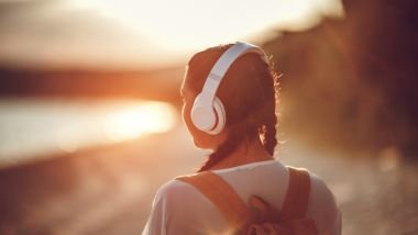 Imagem de uma moça caminhando à beira mar, usando um fone de ouvido branco. Ela está ouvindo a sua música preferida.