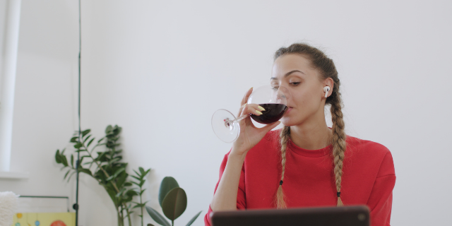 Kaponia Aliaksei / Canva Mulher toma vinho enquanto observa a tela de um computador.