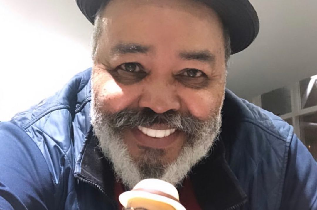 Homem branco de barba grisalha, usando chapéu preto, com expressão sorridente.