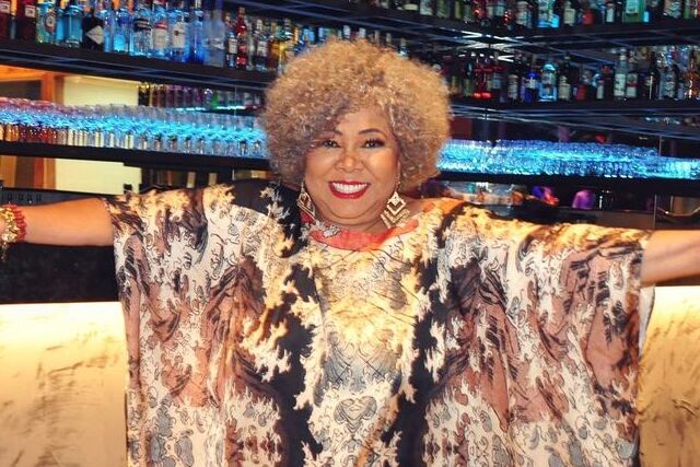 Mulher negra de cabelos curtos e loiros, usando vestido colorido e de braços abertos.