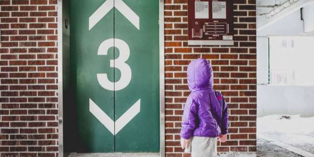 Número três na porta do elevador e criança de casaco roxo olhando para ela