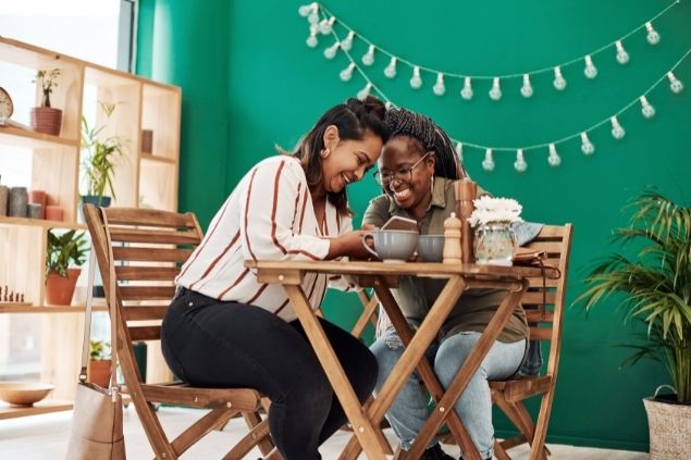 Duas mulheres sentadas em um café rinod enquanto uma apresenta alguém no celular para a outra