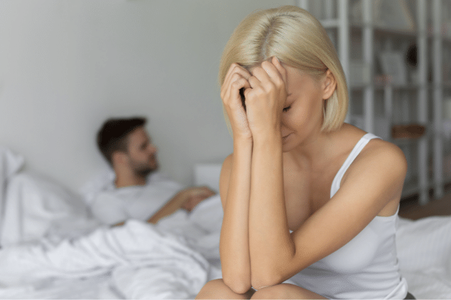 Casal sentado na cama, distantes. A mulher se encontra cabisbaixa. 