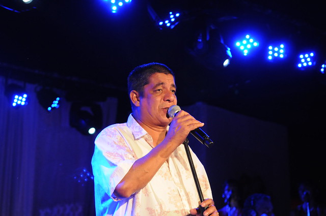 Homem branco de cabelos ralos e pretos, cantando num microfone.