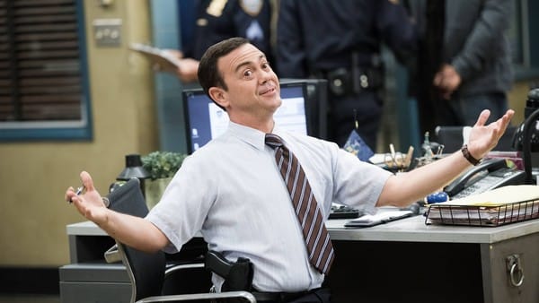 Homem branco de cabelos curtos e pretos, usando roupas sociais, sentado numa cadeira com braços abertos.