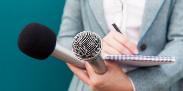 Repórter segurando dois microfones e um bloco de notas