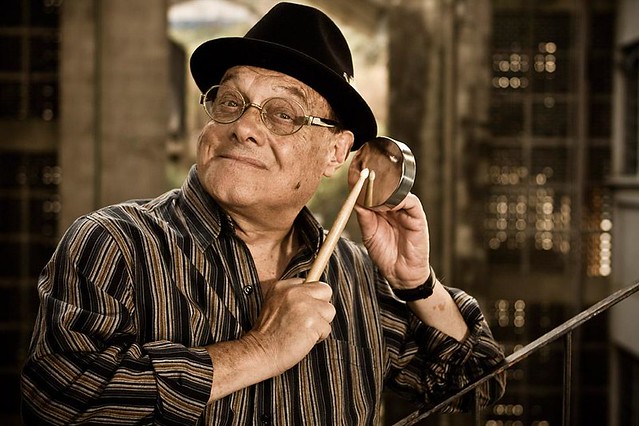 Idoso branco usando chapéu preto e óculos, segurando pequeno instrumento.