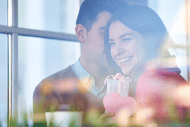 Homem sussurando elogios no ouvido de uma mulher, enquanto lhe entrega presentes. 