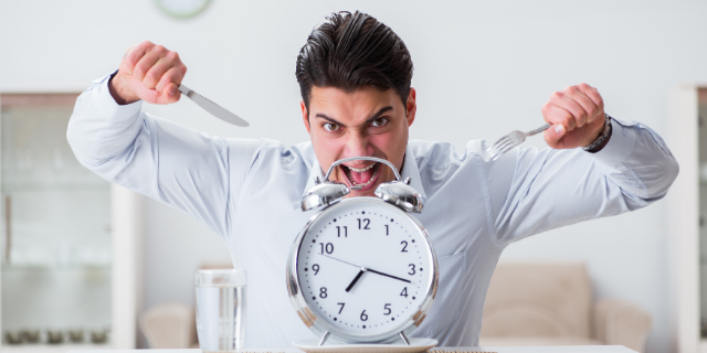 Homem com semblante raivoso segura um garfo e uma faca em cada mão, apontando-os para um relógio.