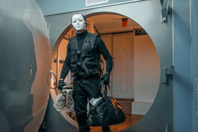 Ladrão vestindo máscara enquanto saí de cofre de banco com malas de dinheiro.