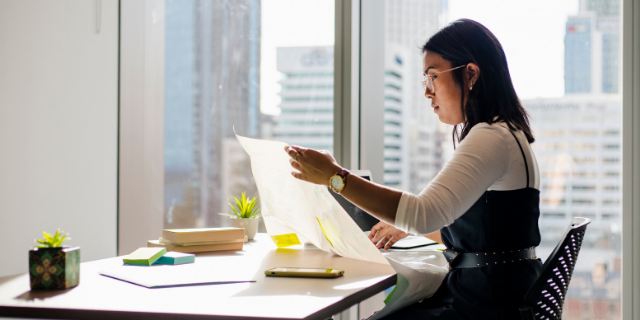 Mulher sentada à mesa observa papéis.
