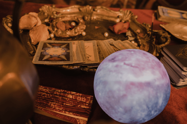 Há várias cartas de Tarot viradas para baixo em cima de um prato sobre uma mesa. Ao lado, há uma bola de cristal e outros objetos.
