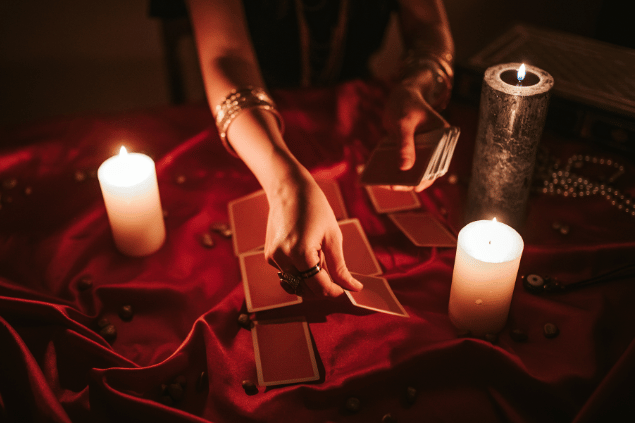 Urbazon / Getty Images Signature / Canva As mãos de uma taróloga segurando e posicionando cartas de Tarot viradas para baixo estão em destaque na imagem. Há velas ao lado.
