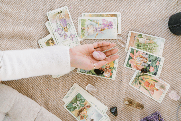 A mão de uma taróloga segurando uma pedra está em destaque na imagem. Embaixo, há algumas cartas de Tarot espalhadas.