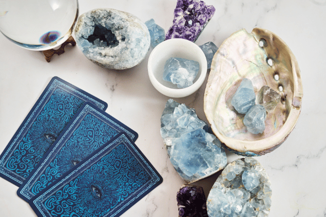 Algumas cartas de Tarot viradas para baixo estão em destaque na imagem. Ao lado, há cristais e outros objetos.
