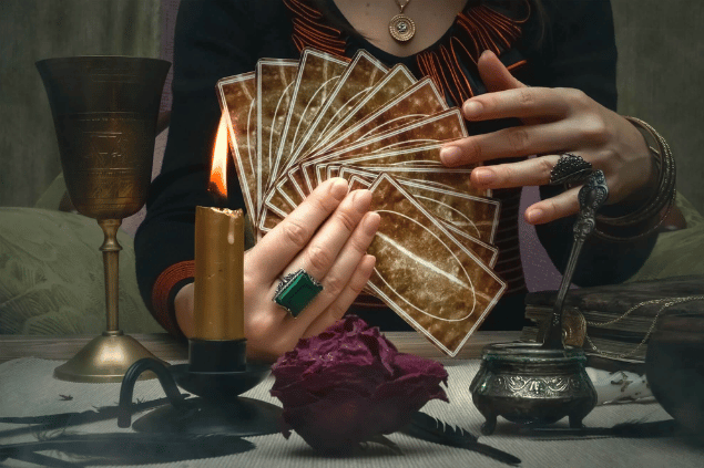 As mãos de uma taróloga segurando cartas de Tarot estão em destaque na imagem. Ao lado, há uma vela e outros objetos.