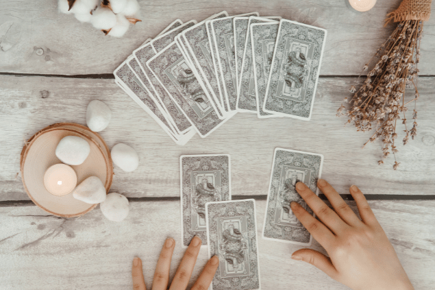 Изображения пользователя annu1tochka / Canva As mãos de uma taróloga posicionando cartas de Tarot viradas para baixo estão em destaque na imagem. Ao lado, há uma vela e outros objetos.