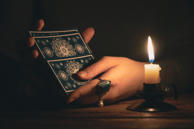 As mãos de uma taróloga segurando cartas de Tarot viradas para baixo estão em destaque na imagem. Há uma vela ao lado.