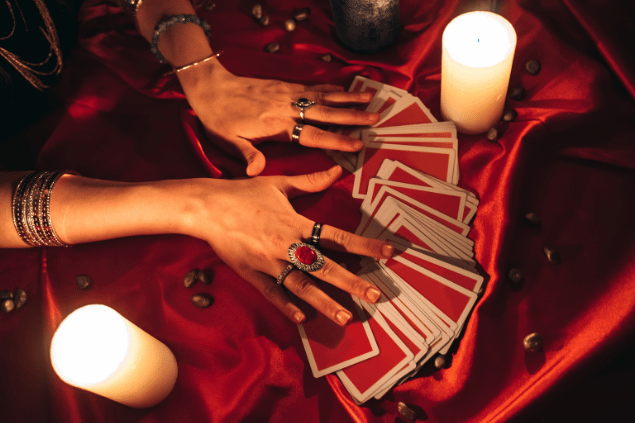 As mãos de uma taróloga sobre várias cartas de Tarot viradas para baixo estão em destaque na imagem. Há velas ao lado.