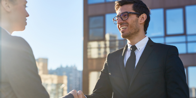 Homem sorri e cumprimenta um à sua frente. Ambos estão vestidos socialmente.