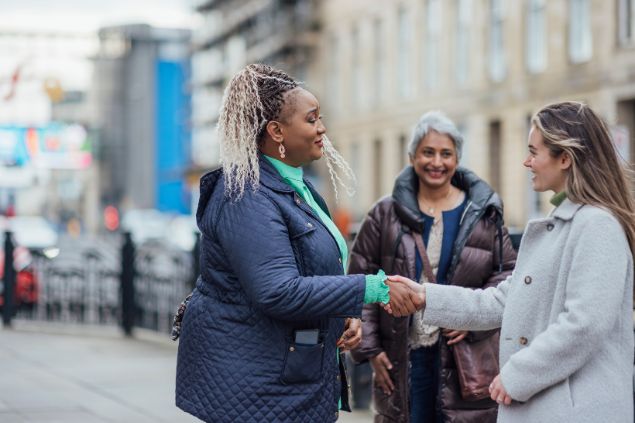 Imagem de três mulheres na rua, e duas delas estão se cumprimentando como se estivessem sendo apresentadas uma pra outra