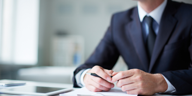Homem trajado socialmente e sentado à mesa. Ele segura uma caneta entre os dedos. 