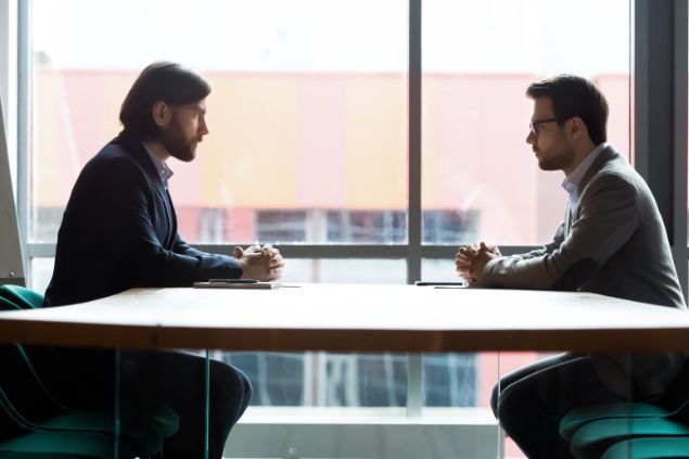 Dois homens sentados à mesa, um de frente para o outro, se encarando.
