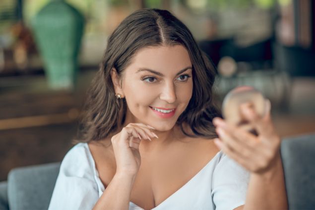 Imagem de uma mulher se olhando em um espelho pequeno e sorrindo